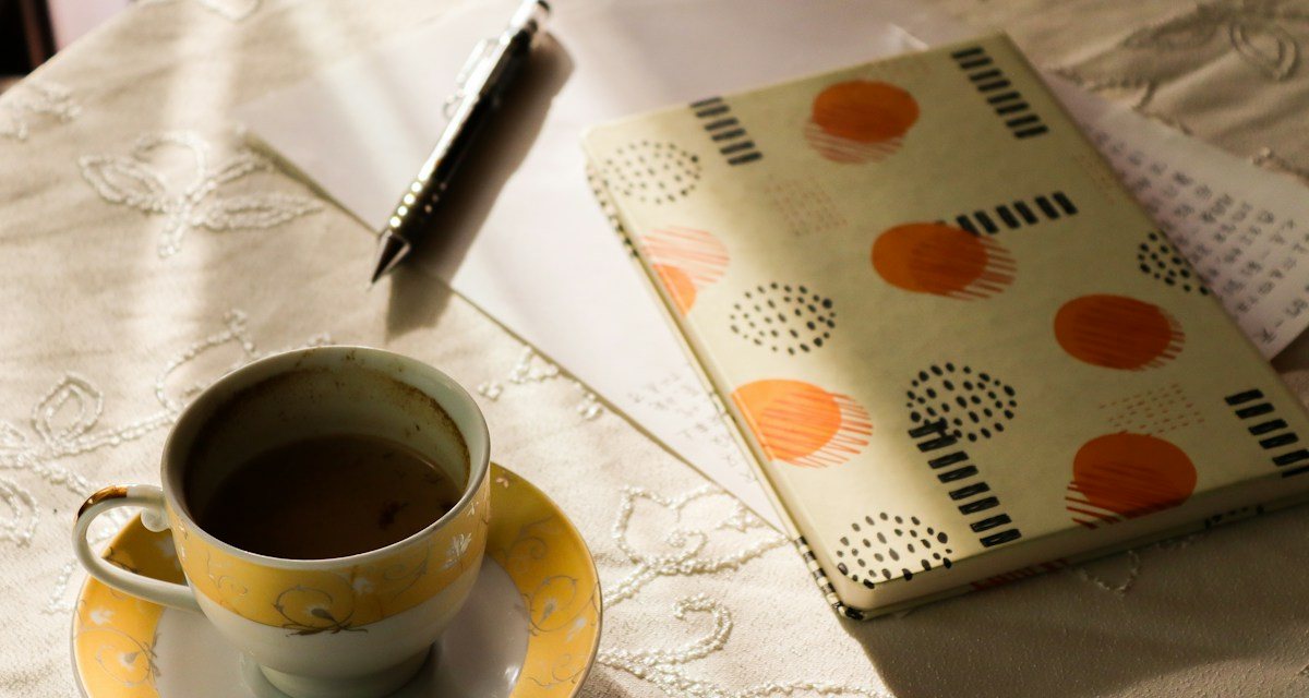 日だまりのテーブルに置かれた、紅茶のカップと柄のノート、ペン